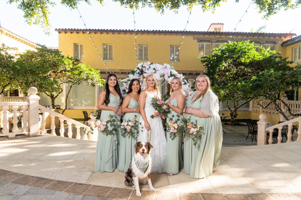 4 bridesmaids with teal dresses and a bride standing with the dog of honor sitting in front of them.